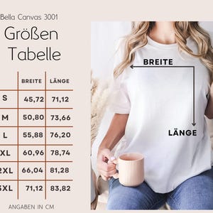 May include: A white Bella Canvas 3001 t-shirt with a size chart in centimeters. The chart shows measurements for width and length, ranging from size S to 3XL. The person is holding a peach-colored mug.