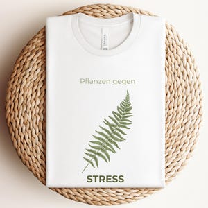 May include: White t-shirt with a crew neck featuring the text "Pflanzen gegen STRESS" and a green fern illustration. The shirt is folded and placed on a woven, circular surface.