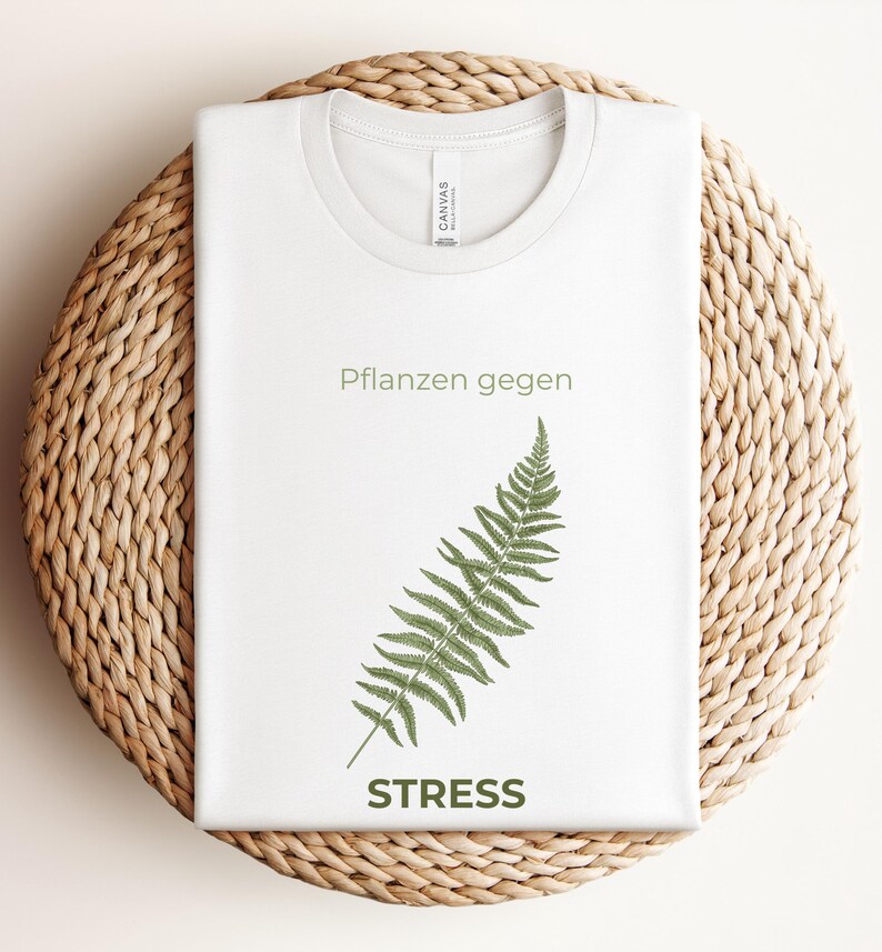 May include: White t-shirt with a crew neck featuring the text "Pflanzen gegen STRESS" and a green fern illustration. The shirt is folded and placed on a woven, circular surface.