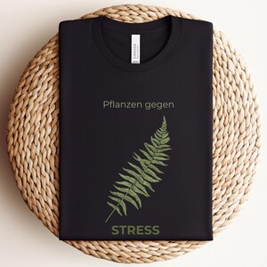 May include: Black t-shirt with the text "Pflanzen gegen STRESS" and a green fern illustration. The shirt is folded on a woven, circular surface. The text and fern are in a light green color.
