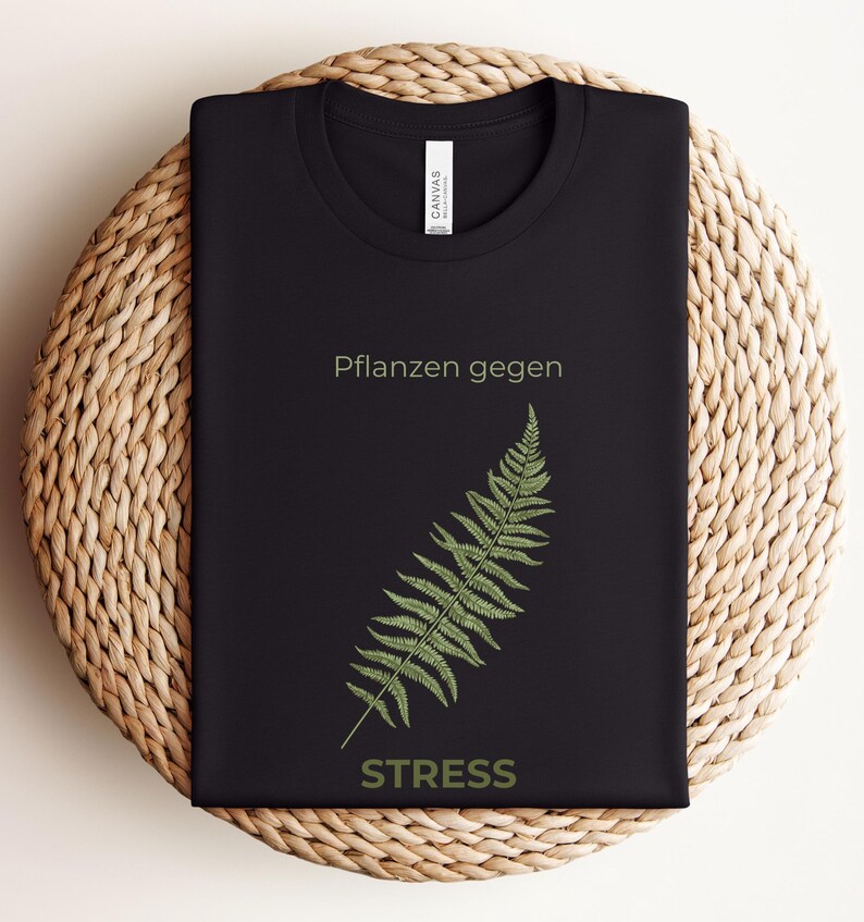 May include: Black t-shirt with the text "Pflanzen gegen STRESS" and a green fern illustration. The shirt is folded on a woven, circular surface. The text and fern are in a light green color.