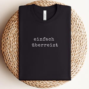 May include: Black t-shirt with the German phrase "einfach &uuml;berreizt" printed in white. The shirt is folded and placed on a woven, circular surface.