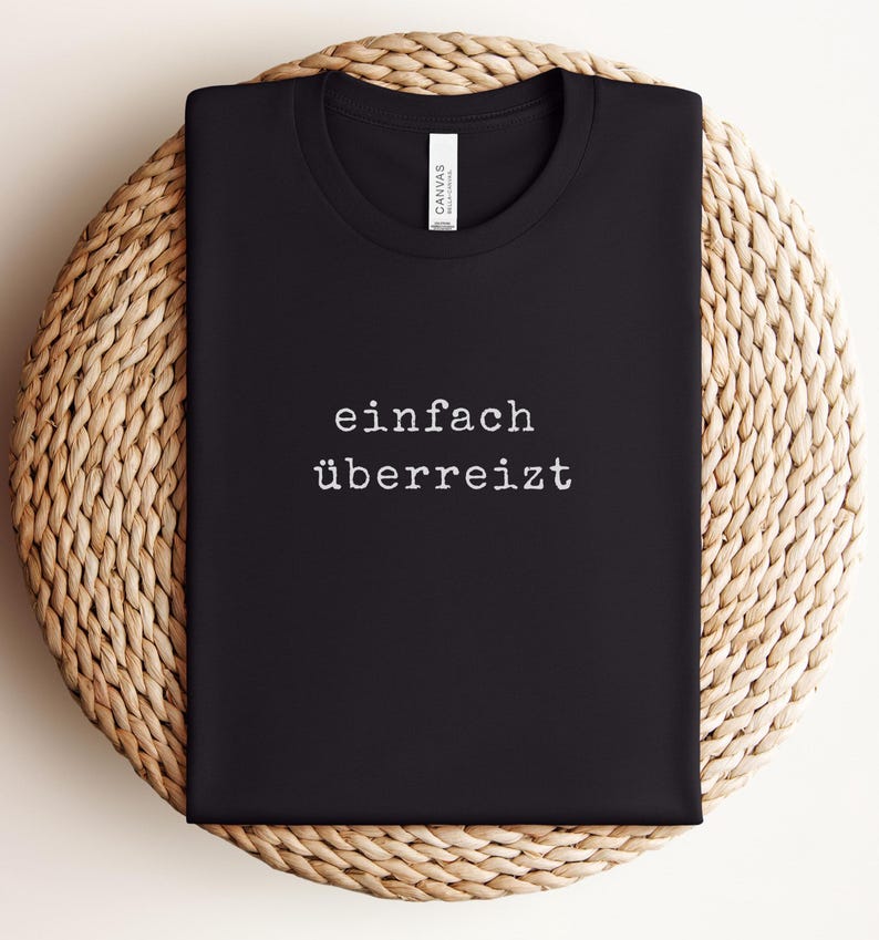 May include: Black t-shirt with the German phrase "einfach &uuml;berreizt" printed in white. The shirt is folded and placed on a woven, circular surface.