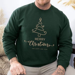 May include: Dark green sweatshirt with a gold-colored Christmas tree design and the words "MERRY CHRISTMAS AND HAPPY NEW YEAR". The sweatshirt is a crew neck style.
