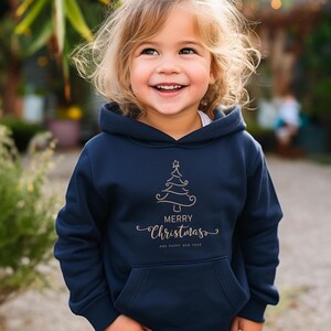 May include: A navy blue hooded sweatshirt with a front pocket. The sweatshirt features a gold-colored Christmas tree design and the words "Merry Christmas" and "And Happy New Year" in a script font.