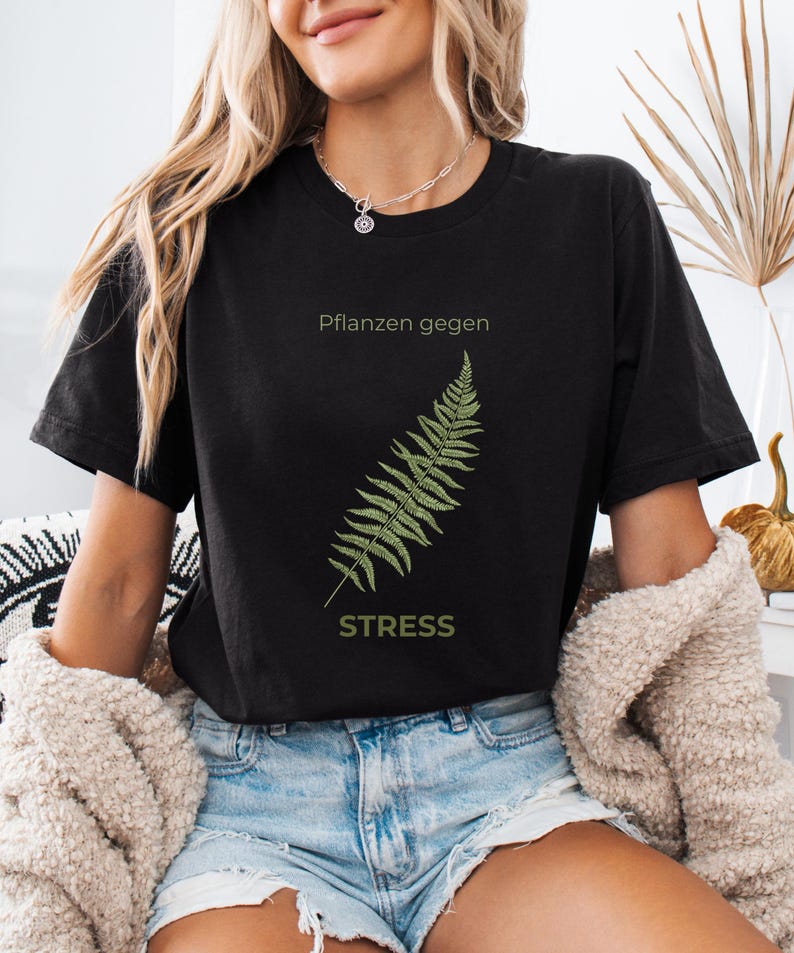 May include: Black t-shirt with a green fern illustration and the text "Pflanzen gegen STRESS". The shirt is made of a soft material and is suitable for casual wear. The design is simple and nature-inspired.