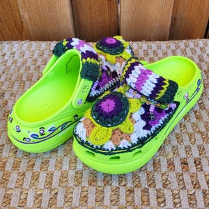 May include: Bright green clogs with decorative crochet accents. The crochet features a floral design with purple, yellow, and white colors. The clogs have a strap and are decorated with painted details.