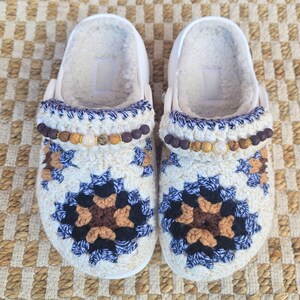 May include: A pair of white clogs with a cream-colored, crocheted top featuring a floral pattern in shades of blue, brown, and black. The clogs have a faux fur lining and are adorned with a beaded accent.