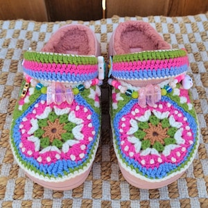 May include: A pair of pink crocheted clogs with colorful yarn and beads. The clogs have a white and green geometric pattern with pink, blue, and brown accents.