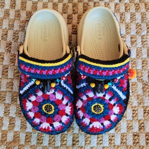 May include: A pair of beige Crocs sandals with intricate, colorful crochet detailing. The crochet design features a floral pattern in shades of blue, red, pink, and white, with yellow and black accents. The sandals have a decorative pom-pom and bead embellishments.