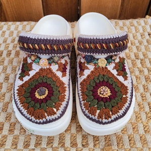 May include: A pair of white clogs with intricate crochet detailing. The crochet design features a floral pattern in shades of brown, green, and burgundy. The upper strap is adorned with a decorative trim and beads, adding a touch of elegance to the footwear.