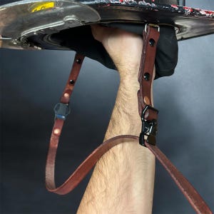 Könnte beinhalten: Ein braunes Lederkameraband mit einem schwarzen Metallclip und verstellbarer Länge. Das Band wird von einer Hand gehalten.