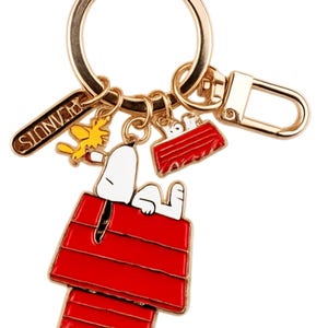 May include: Gold-tone keychain featuring Snoopy resting on his red doghouse, with other Peanuts-themed charms. Includes a charm with the word "PEANUTS" and charms of Woodstock and Snoopy's doghouse.