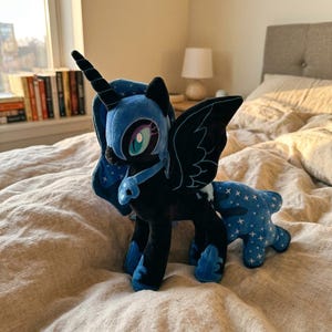 May include: A plush toy of a dark blue and black alicorn with a horn, large wings, and a mane featuring a star pattern. The character has large teal eyes, a blue collar with a crescent moon emblem, and a tail decorated with white stars.