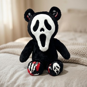 Pode incluir: Um urso de pelúcia com corpo de pelo preto e rosto de máscara branca com uma expressão de boca aberta. O urso tem orelhas arredondadas e pés decorados com padrões vermelhos e brancos, incluindo um pequeno gráfico de máscara no pé direito.