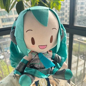 May include: A plush doll featuring teal hair styled in long twin tails, a grey and teal outfit with a matching necktie, and black headphones. The doll has a light skin tone, embroidered brown eyes, and a smiling mouth with pink cheeks.