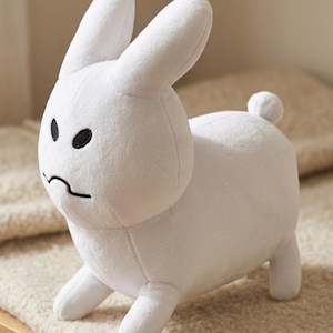 May include: A white plush rabbit toy with long upright ears and a small round tail. The stuffed animal features embroidered black oval eyes and a simple black curved line for a mouth, giving it a distinct, serious facial expression.