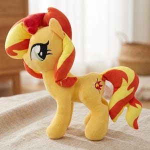 Puede incluir: Un peluche de un poni amarillo con crin y cola de color rojo y amarillo. El poni tiene grandes ojos verdes y un símbolo de sol rojo en el flanco. La figura se mantiene en pie sobre cuatro patas y presenta una construcción de tela suave.