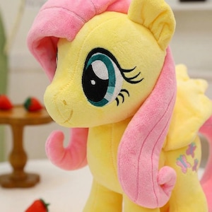 Può includere: Un peluche a forma di pony giallo dalla consistenza morbida, dotato di una lunga criniera e coda rosa. Il pony presenta grandi occhi ricamati color verde acqua con ciglia nere e un piccolo sorriso. Tre piccole farfalle rosa e blu sul fianco.
