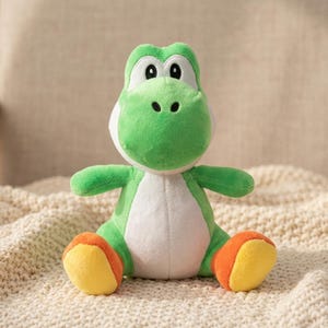 May include: A plush toy shaped like a green dinosaur with a white belly, large oval eyes, and rounded snout. The creature has bright yellow feet with orange accents on the ankles, featuring soft fabric construction and a seated posture.