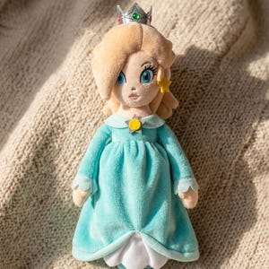 May include: A plush doll of a character with blonde hair, wearing a silver crown with red and green gems, a light blue dress with a white underskirt, and a yellow star brooch at the neckline. The doll has blue eyes and a small yellow star earring.