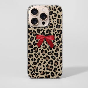 May include: A phone case with a leopard print pattern and a red bow. The case has a clear edge and a camera aperture. The leopard print has black spots on a beige background.