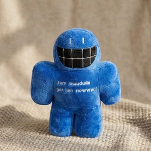 Puede incluir: Un peluche azul con forma de figura geométrica, con una boca ancha negra de patrón cuadriculado y dos ojos blancos pequeños. El pecho presenta un texto bordado en blanco que dice: tem bluudude get jnn nowwwll.