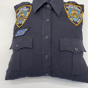 May include: A dark blue pillow shaped like a police uniform shirt with two embroidered patches that say "Police Department City of New York".