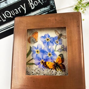 May include: A square, copper-colored box with a glass window displaying a floral and butterfly design. The design features blue flowers, orange and black butterflies, and a bee. The box has a decorative border and the text "LIQUARY BOXES" is visible in the background.