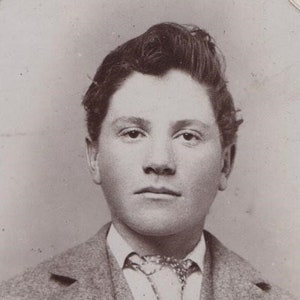 May include: A black and white portrait of a young man with a serious expression. He is wearing a suit jacket with a patterned tie. The photo is faded and has a vintage look.