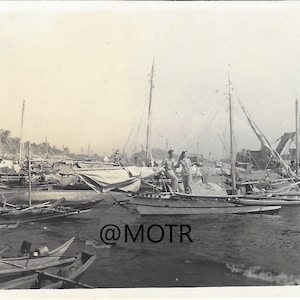 May include: Black and white photograph of a harbor scene filled with numerous small boats. Two people stand on a boat in the foreground. The image has a vintage aesthetic, with the text "@MOTR" in the lower right corner.