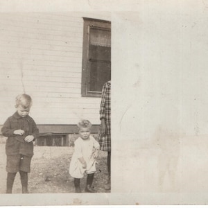 May include: Vintage black and white photograph featuring two young children standing in front of a house. One child wears a dark jacket and shorts, the other a light dress. A person in a plaid shirt stands nearby.