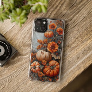 May include: Clear phone case featuring a fall-themed design with pumpkins, orange flowers, and autumn leaves. The case is on a wooden surface, with a camera and greenery in the background. The design has a watercolor effect.