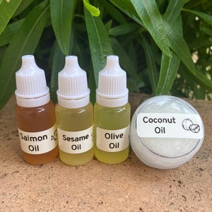 May include: Three small plastic dropper bottles containing amber, pale yellow, and light yellow oils labeled Salmon Oil, Sesame Oil, and Olive Oil, alongside a small round clear container filled with white Coconut Oil with a coconut graphic.