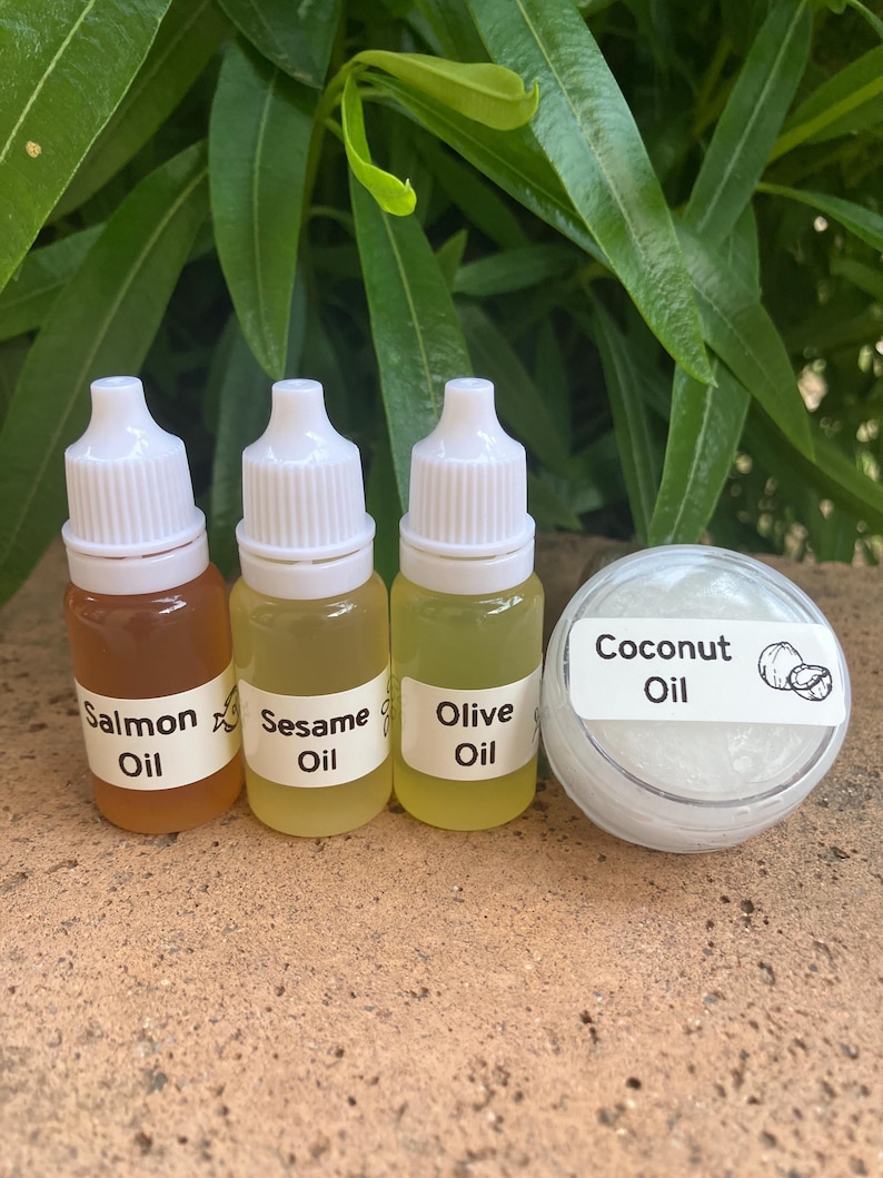 May include: Three small plastic dropper bottles containing amber, pale yellow, and light yellow oils labeled Salmon Oil, Sesame Oil, and Olive Oil, alongside a small round clear container filled with white Coconut Oil with a coconut graphic.