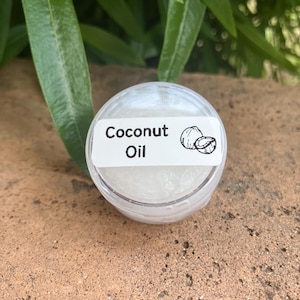 May include: A small, round, clear plastic container filled with a white, semi-solid substance. A white rectangular label is affixed to the lid, featuring the black text "Coconut Oil" and a small line drawing of a coconut.