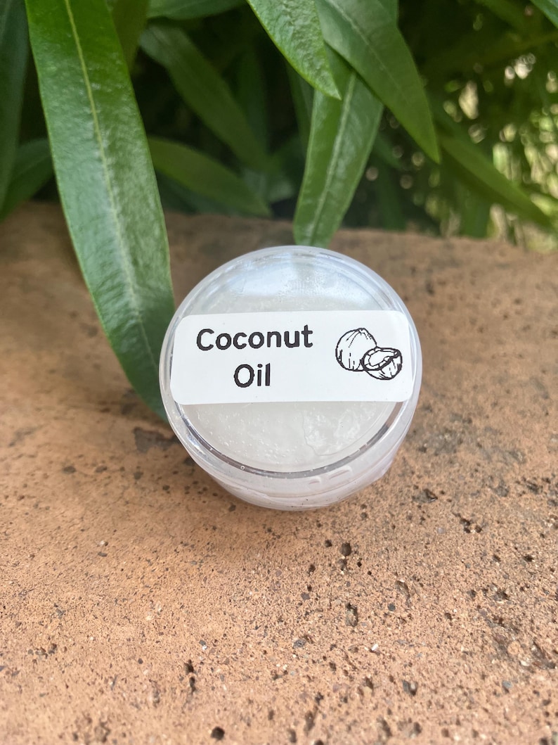 May include: A small, round, clear plastic container filled with a white, semi-solid substance. A white rectangular label is affixed to the lid, featuring the black text "Coconut Oil" and a small line drawing of a coconut.