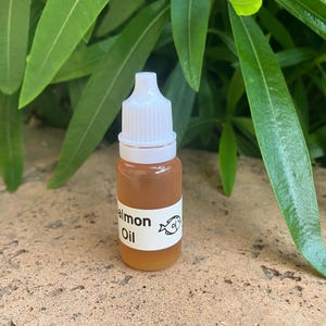 May include: A small, clear plastic dropper bottle filled with an amber-colored liquid. The bottle features a white label with the text "almon Oil" and a small graphic of a fish. It has a white, ribbed, screw-on cap with a pointed tip.