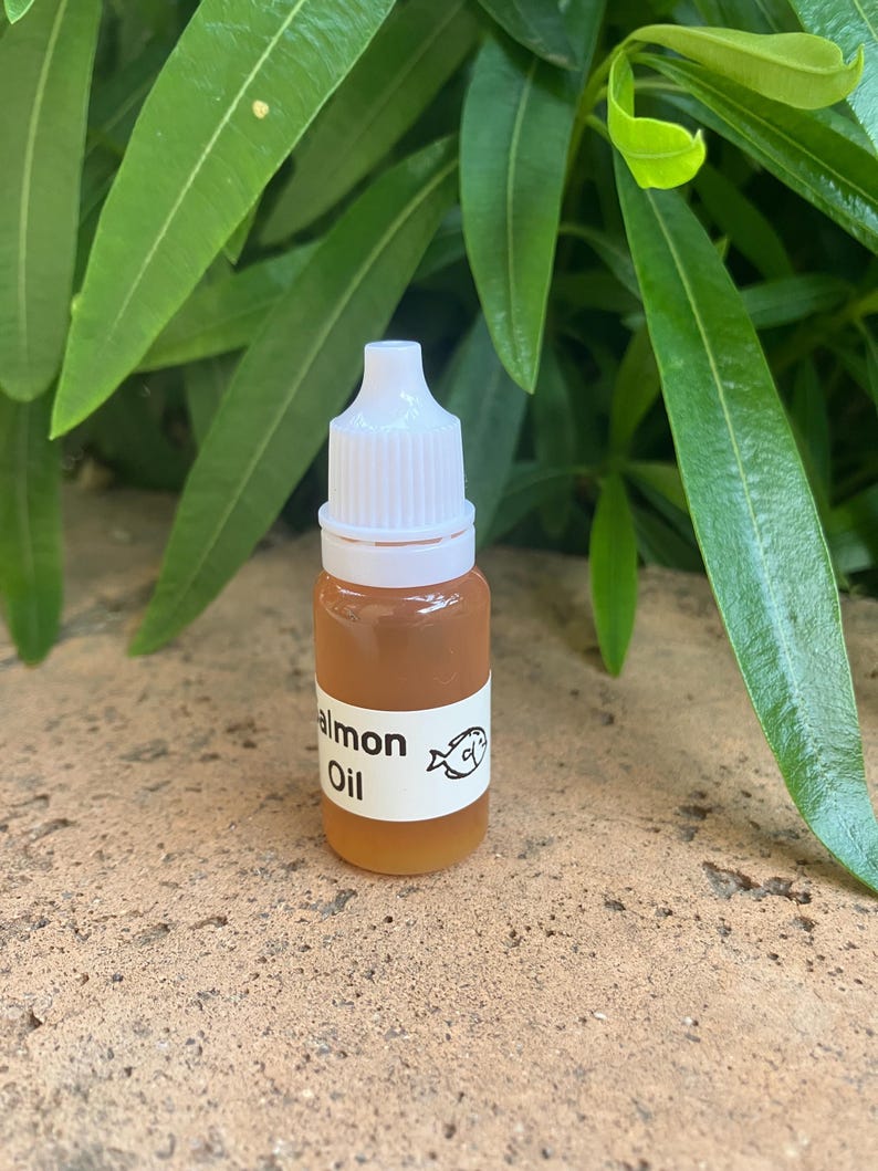 May include: A small, clear plastic dropper bottle filled with an amber-colored liquid. The bottle features a white label with the text "almon Oil" and a small graphic of a fish. It has a white, ribbed, screw-on cap with a pointed tip.