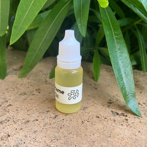 May include: A small, clear plastic dropper bottle filled with a pale yellow liquid. The bottle features a white label with the text "Sesame Oil" printed in black, accompanied by a simple graphic illustration of several small sesame seeds.