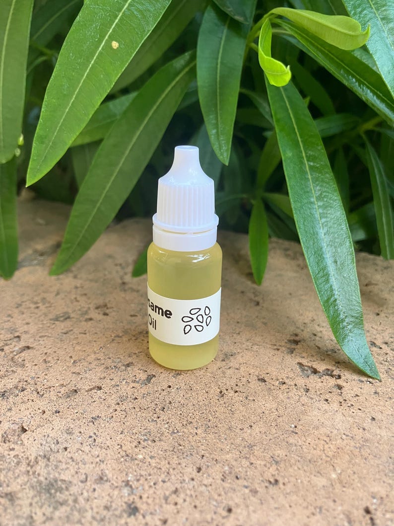 May include: A small, clear plastic dropper bottle filled with a pale yellow liquid. The bottle features a white label with the text "Sesame Oil" printed in black, accompanied by a simple graphic illustration of several small sesame seeds.