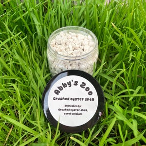 May include: A clear glass jar filled with small, beige crushed oyster shells. The jar sits on a bed of green grass, with a black and white label that reads "Abby's Zoo Crushed oyster shell" and lists the ingredients.