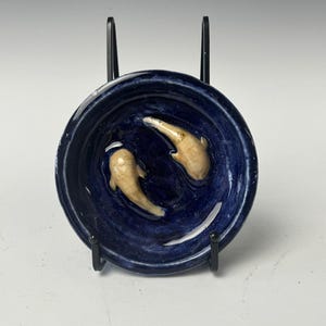 May include: A small blue ceramic dish with two white fish sculpted into the surface. The dish is on a black stand.