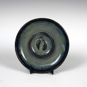 May include: A small, round, blue and white ceramic plate with a raised design in the center. The plate has a dark blue rim and a light blue center with a raised design of two small, rounded shapes.