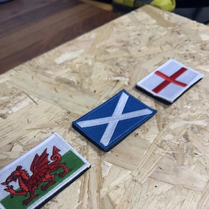 May include: Three rectangular embroidered patches displaying national flags. The patches feature the Welsh flag with a red dragon, the Scottish flag with a white saltire on a blue background, and the English flag with a red cross on a white background.