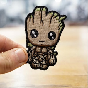 May include: Embroidered patch depicting a cartoon character resembling a tree. The patch has a tan body, black eyes, and green detailing. The character is outlined in black.