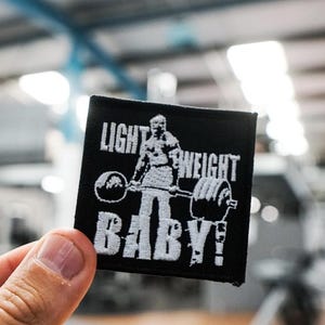 May include: Black square patch with white embroidered text and a weightlifter graphic. The text reads "LIGHT WEIGHT BABY!" The patch features a weightlifter lifting a barbell with weights. The background is a blurred gym setting.