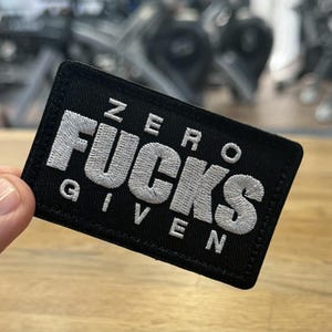 May include: Black rectangular patch with white embroidered text reading "ZERO FUCKS GIVEN". The patch is held up against a wooden surface, with gym equipment visible in the background.
