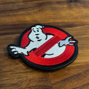 May include: Embroidered patch featuring the Ghostbusters logo. The patch has a white ghost figure inside a red circle with a red slash through it, all outlined in black. The patch is on a wooden surface.