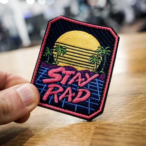 May include: Embroidered patch with a retro design featuring a yellow sun, palm trees, and the words "STAY RAD" in pink. The patch has a black background with a pink border and a blue grid pattern.